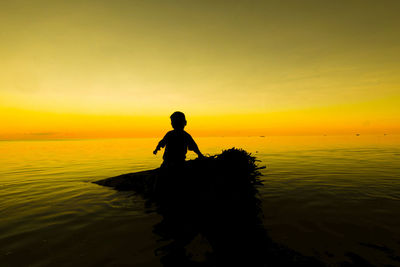 Silhouette boy on fallen tree in sea against sky during sunset