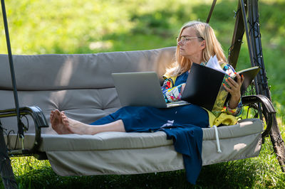 Mature woman with laptop and documents working in garden on rocking couch, green home office concept
