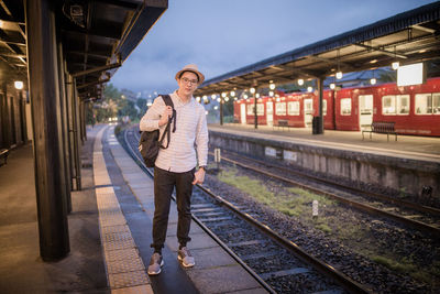 Portrait of mid adult man with backpack standing at railroad station platform against sky