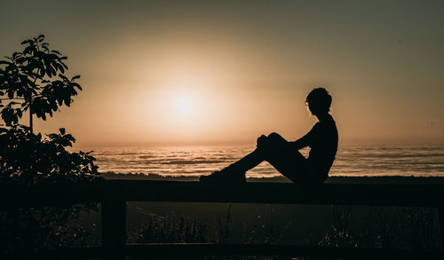 Silhouette woman sitting on railing at beach against sky during sunset