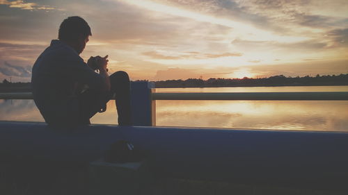 Man photographing while sitting on retaining wall by lake during sunset