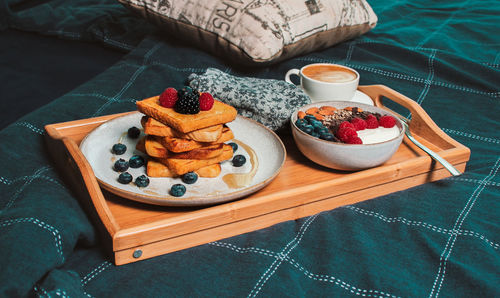 Cozy breakfast in bed, yogurt bowl with berries and nuts, french toasts, coffee cup on wooden plate