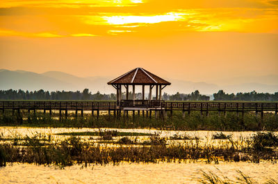 Gazebo by lake against sky during sunset