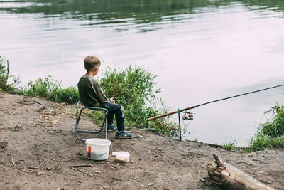 A boy is sitting on the shore of a lake or river with a fishing rod during a family vacation