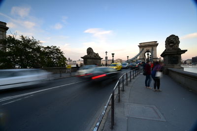 Blurred motion of people and cars on chain bridge against sky