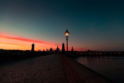 Illuminated street light by silhouette city against sky at sunset