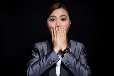 Portrait of surprised businesswoman covering mouth while standing against black background