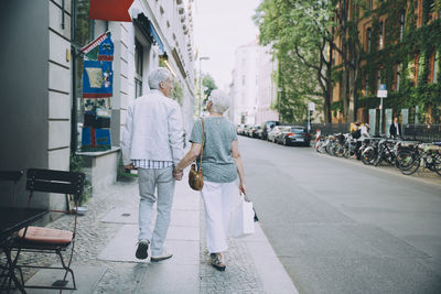 Full length of retired couple holding hands while walking on sidewalk in city