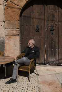 Portrait of adult man sitting on outdoor chair against old wooden door