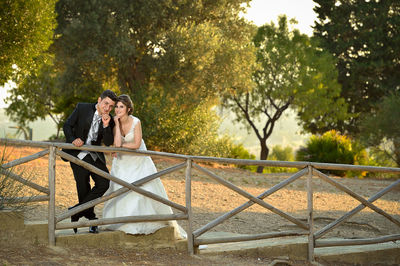 Portrait of wedding couple leaning on railing against trees