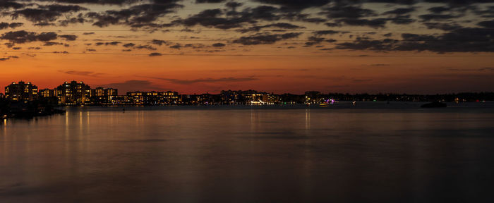 Illuminated buildings by river against sky during sunset