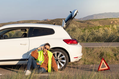 Young man sitting on the road next to a broken car while talking on the phone wating for assistance