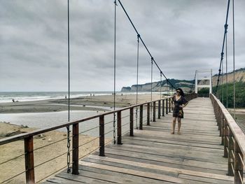 Young woman with hands on hip standing on footbridge against cloudy sky