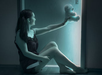 Side view of woman holding teddy bear while sitting by door