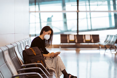Woman wearing mask using mobile phone while sitting at airport