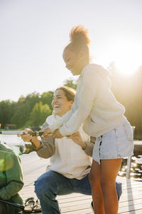 Happy counselor crouching by girl doing fishing while standing on jetty near lake at summer camp