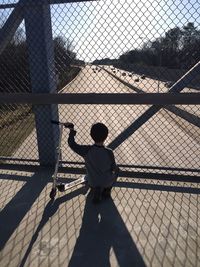 Rear view of boy with push scooter crouching against chainlink fence