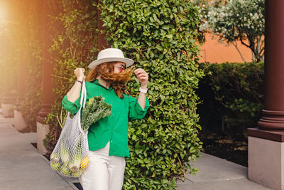 Portrait of a young woman in green shirt holding a mesh bag with veggies on her shoulder 