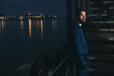 Side view of young man standing at railing on pier over river at night