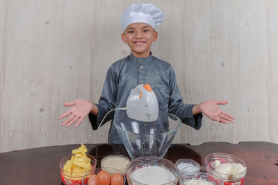 Portrait of boy wearing chef hat with ingredients on table standing against wall at home