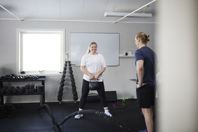 Full length of happy woman exercising with kettlebell in front of fitness instructor at health club