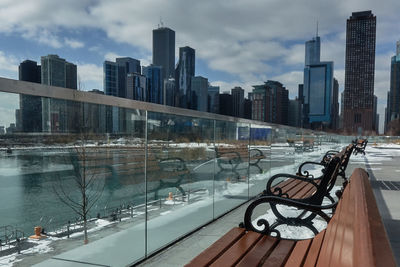 Empty benches by lake against city skyline during winter