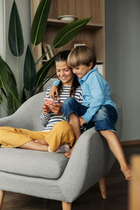 Sister and younger brother are sitting at home in an armchair and playing on the phone