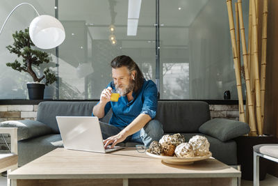 Mature man with a beard drinking orange juice and looking at his lapto