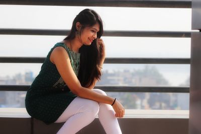 Young woman looking away while sitting on window