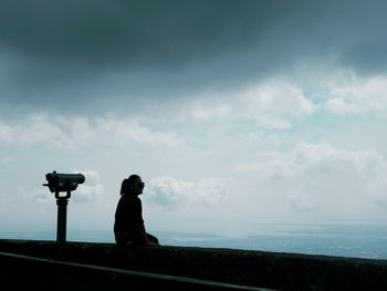 Silhouette of woman sitting at observation point against cloudy sky