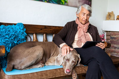 Senior woman using digital tablet with dog sitting on bench at home