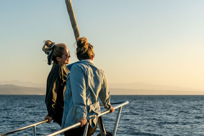 Friends sitting happy on boat bow in sea against clear sky watching sunset looking to horizon.