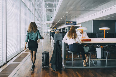 Full length rear view of businesswoman walking with luggage by colleagues waiting at airport departure area