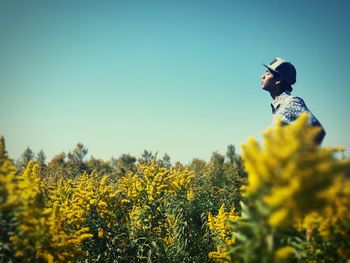 Side view of young man standing amidst yellow flowers on field against clear sky