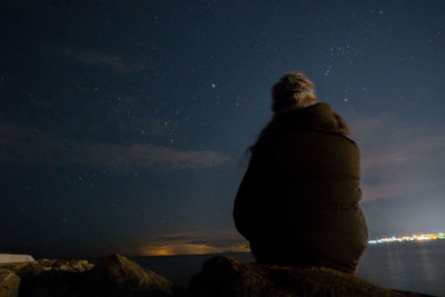 Rear view of man on sitting on cliff by sea against sky at night