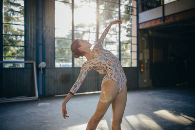 Side view of young graceful ballerina looking up in colorful bodysuit dancing training in light studio