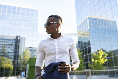 From below stylish african american businessman with jacket on shoulder and in sunglasses walking along street in city and looking away