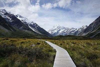 Boardwalk leading towards snowcapped mountains against cloudy sky