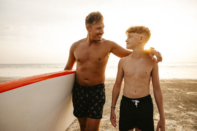 Smiling man holding paddleboard with arm around son at beach during sunset