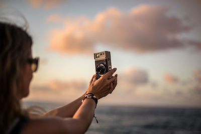 Side view of crop young woman standing at seaside at sunset time and taking picture with analog photo camera