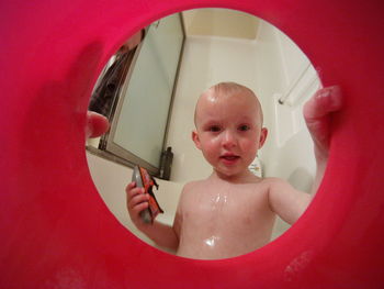 Portrait of shirtless baby boy in bathtub seen through red toy