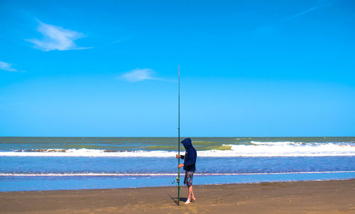 Full length of man holding fishing rod at beach against sky