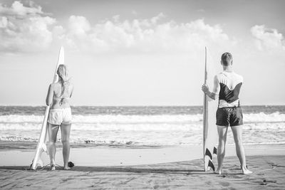 Rear view of people with surfboards standing at beach against sky