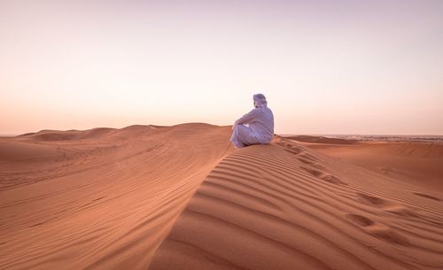 Man sitting on sand dune in desert against clear sky