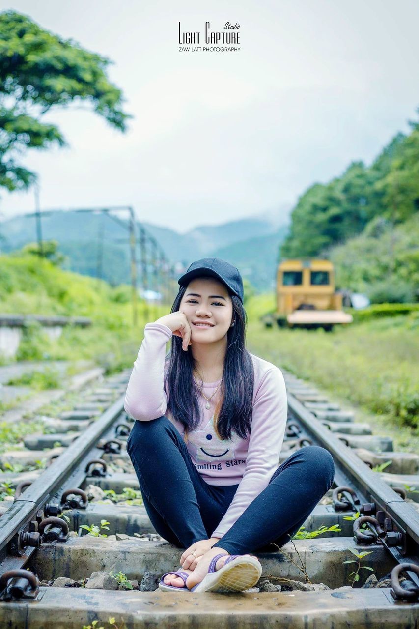 transportation, real people, one person, sitting, railroad track, rail transportation, casual clothing, young adult, day, young women, public transportation, communication, lifestyles, outdoors, sky, technology, nature