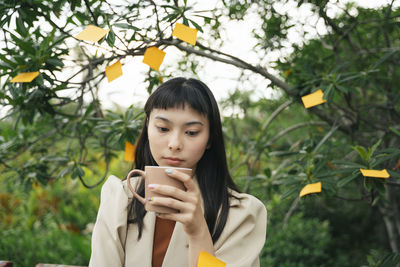 Businesswoman holding coffee cup sitting against tree outdoors