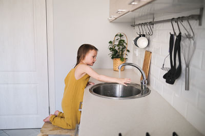 European child of four years old in yellow jumpsuit washes his hands in real bright kitchen