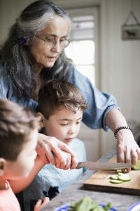 Grandmother assisting grandson cutting cucumber in kitchen at home