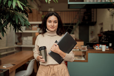 Portrait of businesswoman holding coffee cup and laptop at cafe