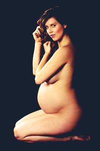 Portrait of naked pregnant young woman covering breasts with hand in hair against black background